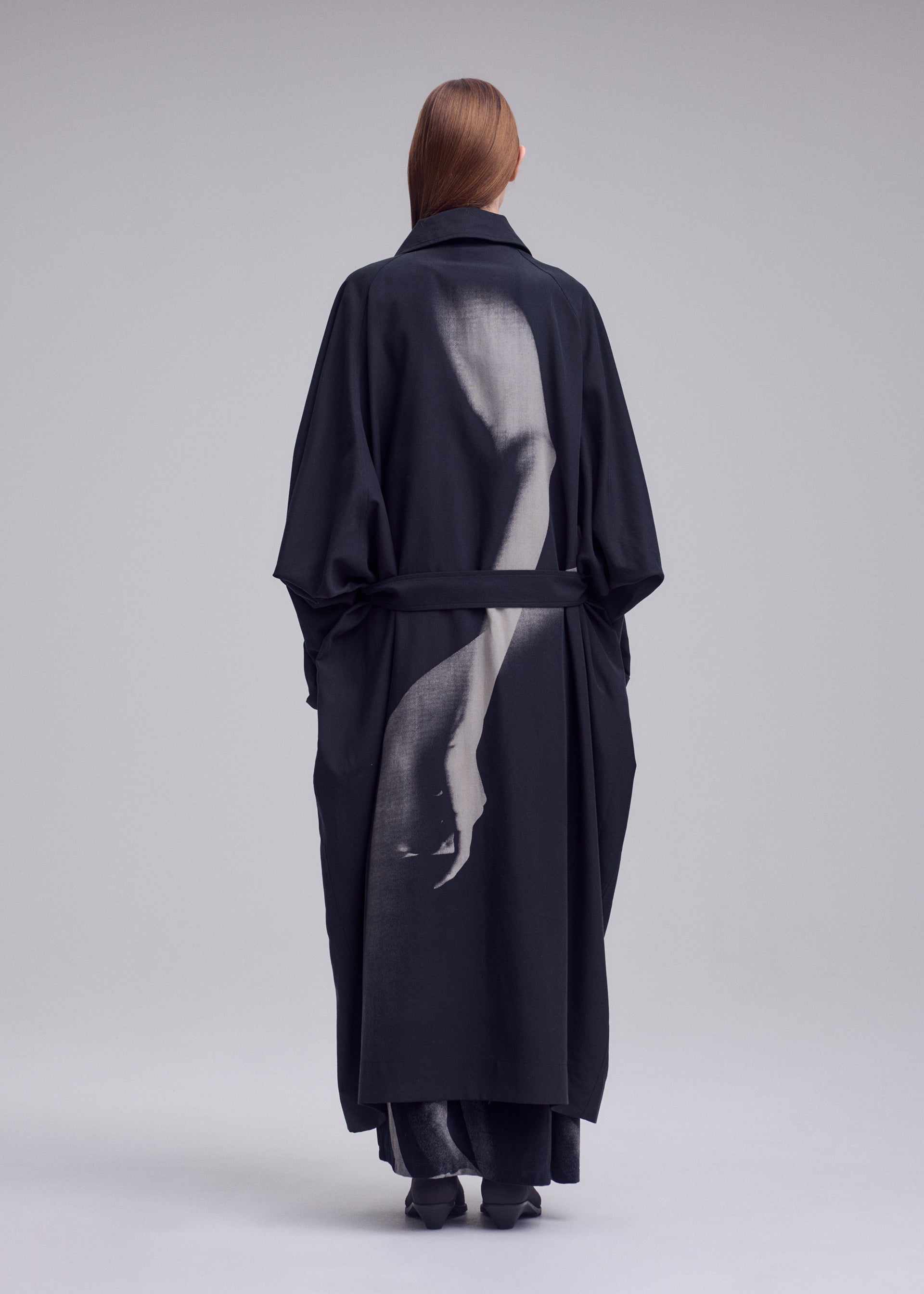 Zoomed view A person wearing the ISSEY MIYAKE “HANDS OF LOVER” coat—crafted from smooth poplin fabric and featuring a grayscale silk-screen discharge print of an arm and hand on the back—stands facing away against a neutral background.