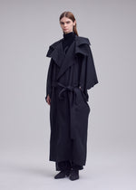 A person with straight brown hair wears the HANDS OF LOVER long, belted black poplin coat by ISSEY MIYAKE, featuring wide lapels and a high collar, standing against a plain light gray background.