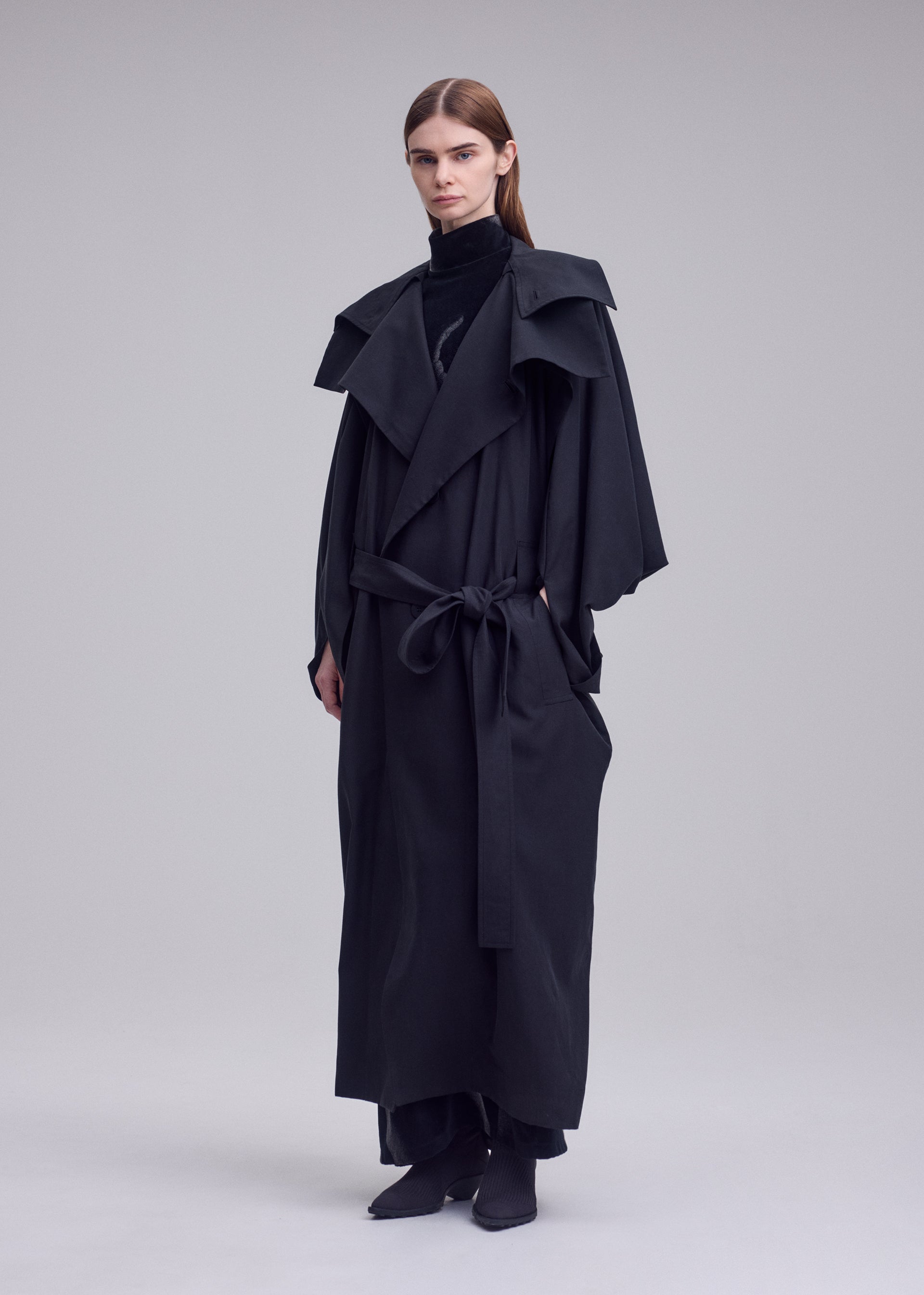 Zoomed view A person with straight brown hair wears the HANDS OF LOVER long, belted black poplin coat by ISSEY MIYAKE, featuring wide lapels and a high collar, standing against a plain light gray background.