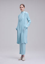 A person wears the ISSEY MIYAKE HATCHING PLEATS light blue pleated coat and matching pants with beige shoes, set against a plain, light gray background.