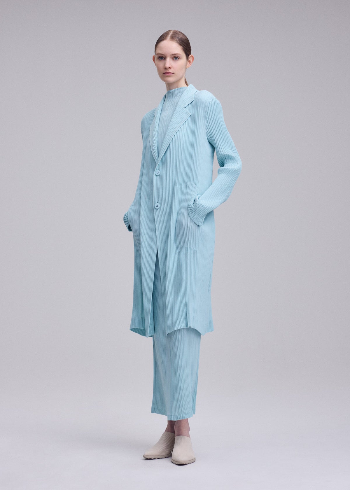 A person models the ISSEY MIYAKE HATCHING PLEATS light blue pleated coat and matching pants with hands in pockets and beige shoes, posed against a plain light gray background. view 5 of 5