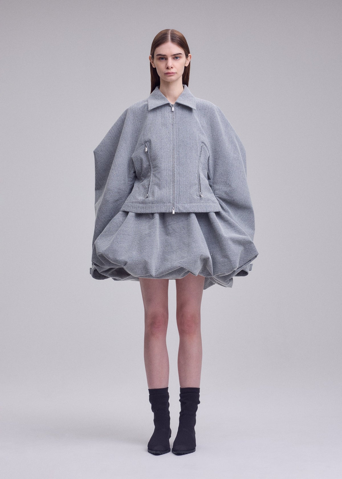 A person stands against a plain background wearing the REFLECTION jacket and matching puffy skirt by ISSEY MIYAKE, featuring a light gray, reflective finish with wide, rounded sleeves, paired with black socks and pointed shoes. view 4 of 7
