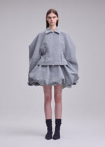 A person stands against a plain background wearing the REFLECTION jacket and matching puffy skirt by ISSEY MIYAKE, featuring a light gray, reflective finish with wide, rounded sleeves, paired with black socks and pointed shoes.