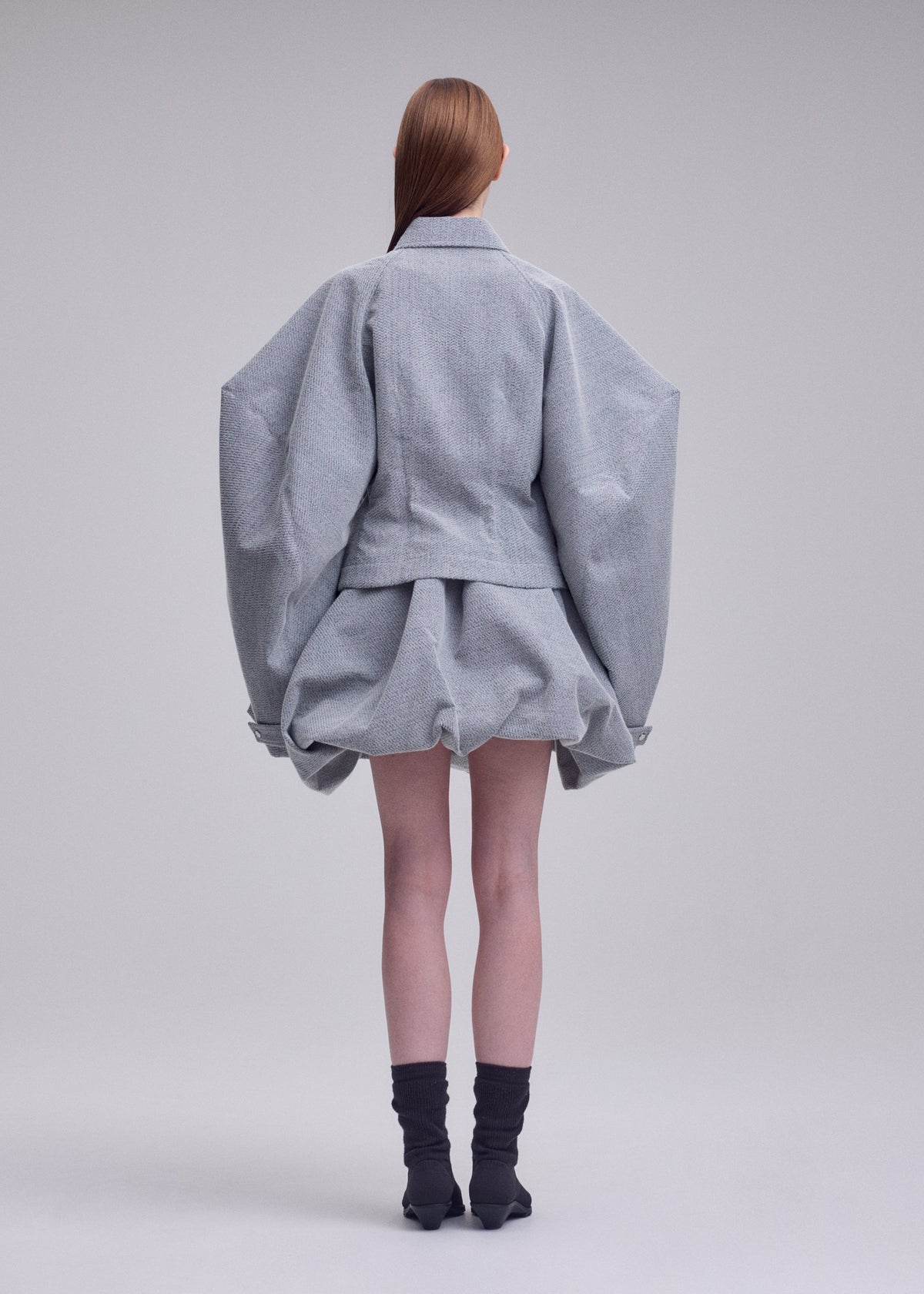A person stands facing away against a plain background, dressed in the ISSEY MIYAKE REFLECTION ensemble—a light gray, reflective jacket with wide sleeves paired with a matching short, puffy skirt. They have long brown hair and wear black socks with black shoes. view 5 of 7