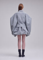 A person stands facing away against a plain background, dressed in the ISSEY MIYAKE REFLECTION ensemble—a light gray, reflective jacket with wide sleeves paired with a matching short, puffy skirt. They have long brown hair and wear black socks with black shoes.