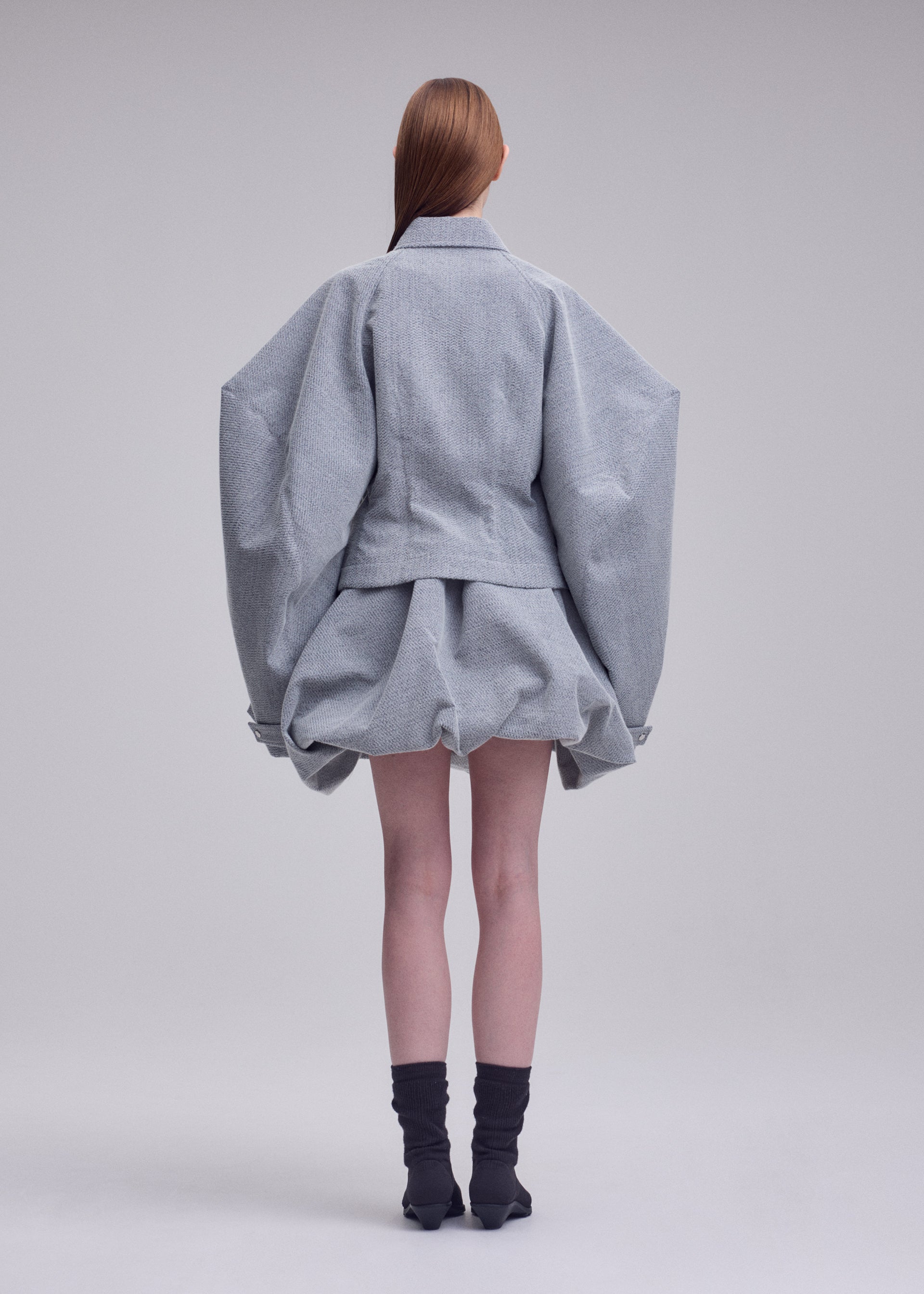 Zoomed view A person stands facing away against a plain background, dressed in the ISSEY MIYAKE REFLECTION ensemble—a light gray, reflective jacket with wide sleeves paired with a matching short, puffy skirt. They have long brown hair and wear black socks with black shoes.