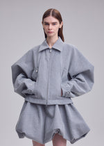 A woman with straight brown hair wears REFLECTION by ISSEY MIYAKE—a loose-fitting, reflective gray jacket with oversized sleeves paired with a matching short, puffy skirt—standing against a plain light background.