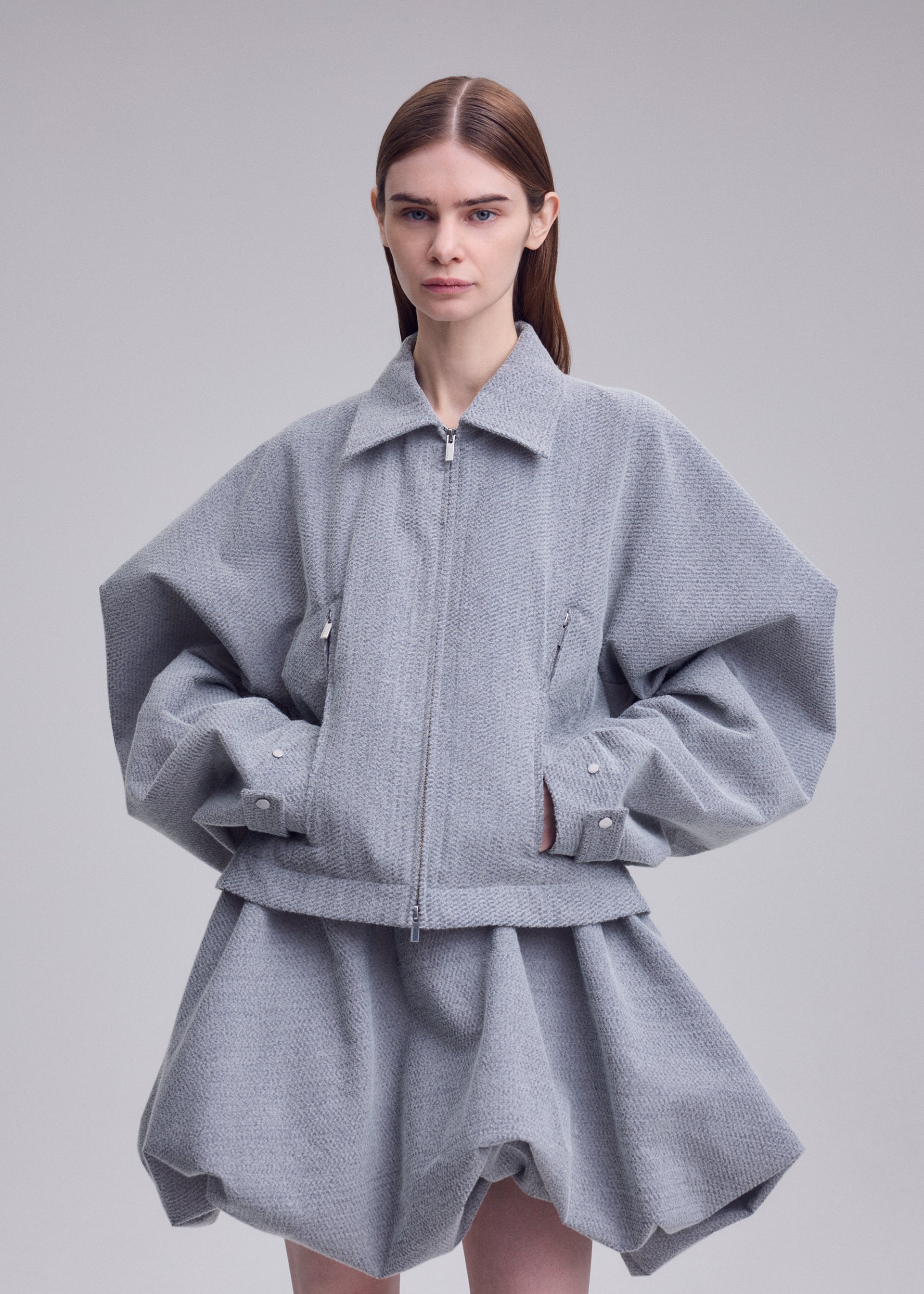 Zoomed view A woman with straight brown hair wears REFLECTION by ISSEY MIYAKE—a loose-fitting, reflective gray jacket with oversized sleeves paired with a matching short, puffy skirt—standing against a plain light background.
