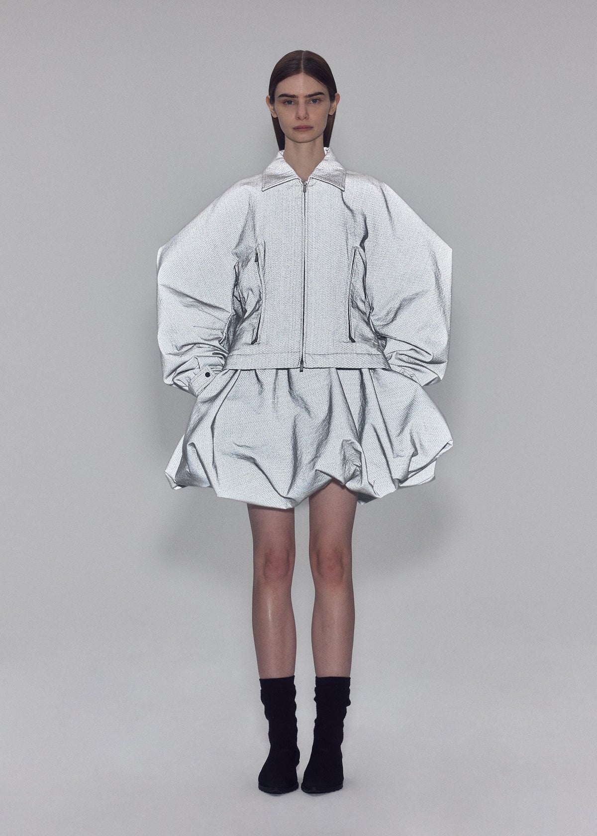 Clad in ISSEY MIYAKE’s REFLECTION ensemble, a person stands against a plain background wearing a reflective silver zip-up jacket featuring voluminous sleeves and a matching puffy skirt, paired with black socks. view 7 of 7