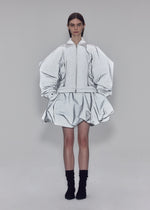 Clad in ISSEY MIYAKE’s REFLECTION ensemble, a person stands against a plain background wearing a reflective silver zip-up jacket featuring voluminous sleeves and a matching puffy skirt, paired with black socks.