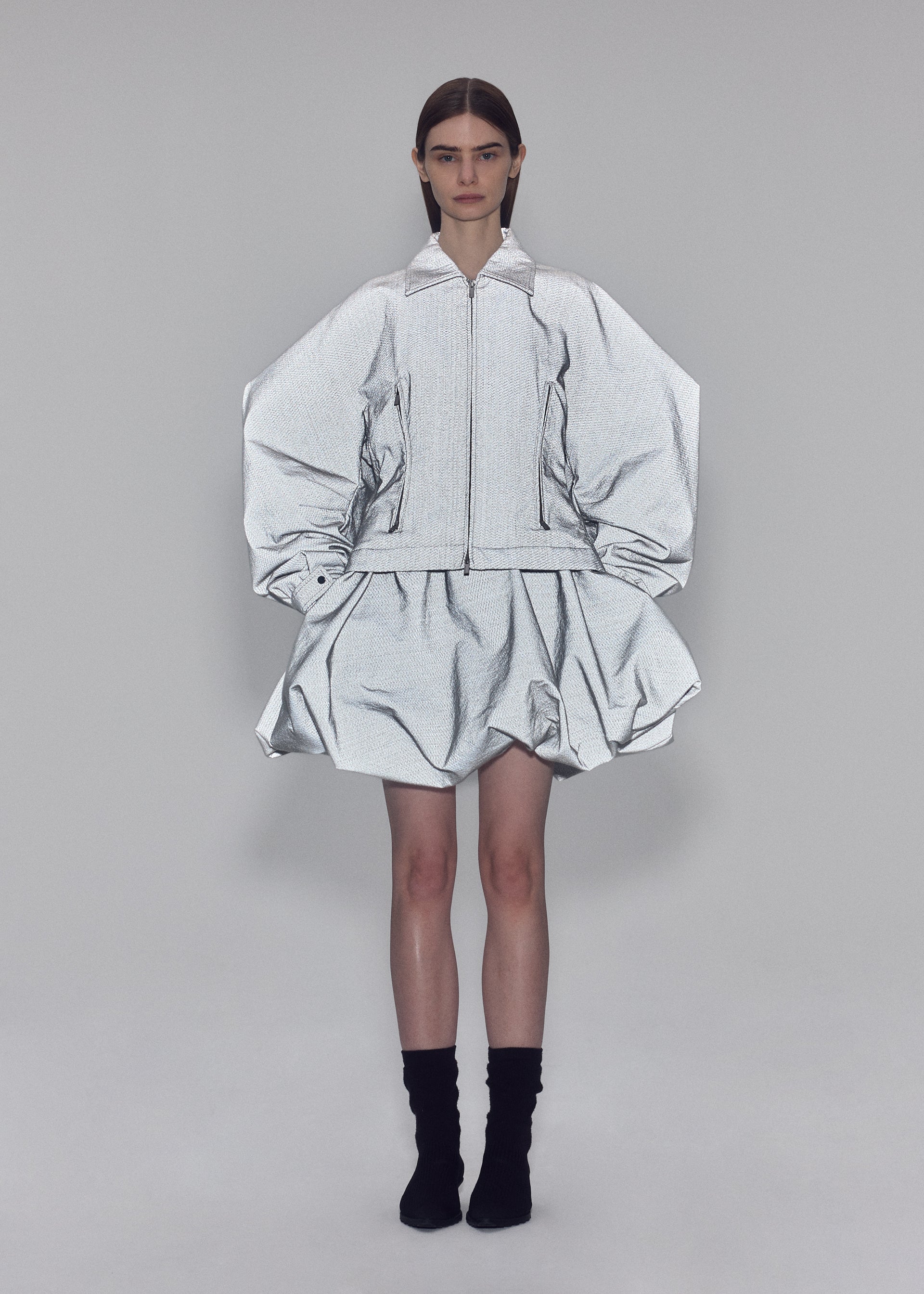 Zoomed view Clad in ISSEY MIYAKE’s REFLECTION ensemble, a person stands against a plain background wearing a reflective silver zip-up jacket featuring voluminous sleeves and a matching puffy skirt, paired with black socks.