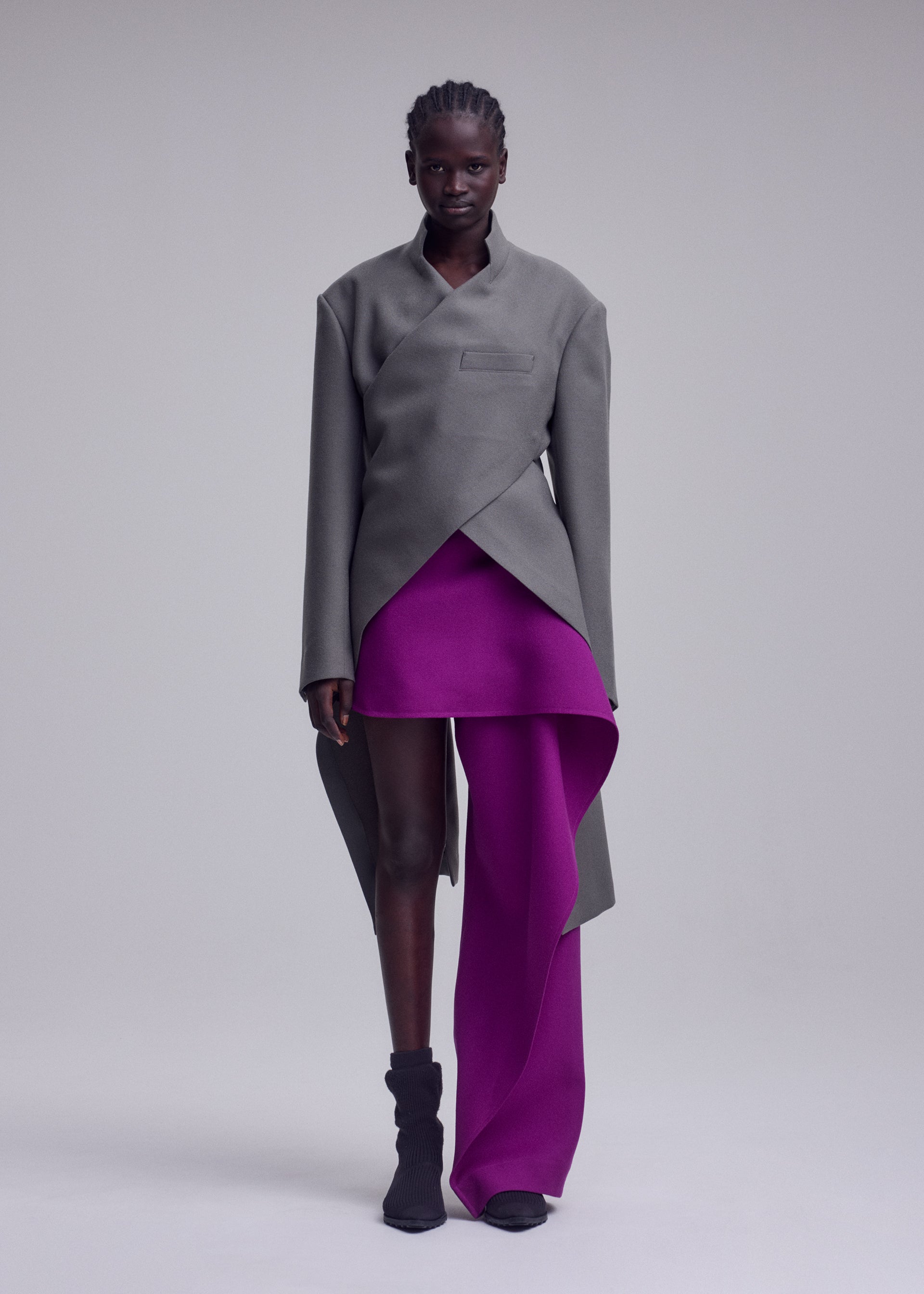 Zoomed view A model wears the ISSEY MIYAKE CALLIGRAPH jacket from the Autumn Winter Collection, featuring an asymmetrical silhouette in gray crafted from premium wool and woven nashiji pebbled fabric. She styles it with a vibrant magenta skirt that boasts a dramatic side drape, and completes the look with black ankle boots against a minimalist light background.