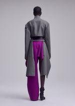 A person stands facing away, wearing the ISSEY MIYAKE CALLIGRAPH gray long coat over a purple skirt and pants. They have braided hair, gray socks, and are set against a plain light gray background.