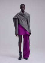 A person stands against a plain light background, wearing a sophisticated ensemble featuring ISSEY MIYAKE’s CALLIGRAPH collection: an asymmetrical gray wrap top, a short magenta skirt layered over matching magenta pants, and a premium wool jacket inspired by the brand’s Autumn Winter Collection. Their look is completed with short, styled hair and black ankle boots.