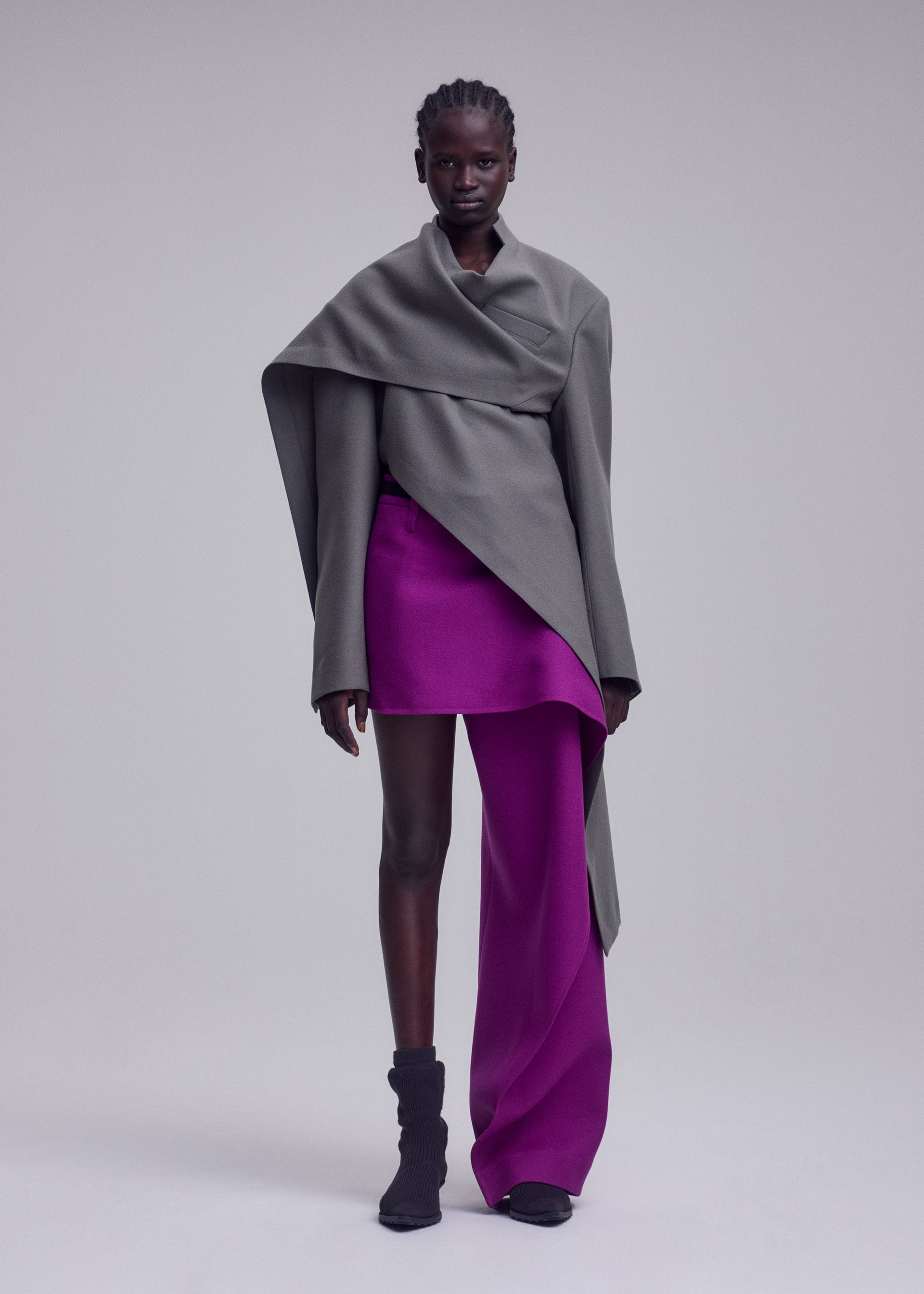 Zoomed view A person stands against a plain light background, wearing a sophisticated ensemble featuring ISSEY MIYAKE’s CALLIGRAPH collection: an asymmetrical gray wrap top, a short magenta skirt layered over matching magenta pants, and a premium wool jacket inspired by the brand’s Autumn Winter Collection. Their look is completed with short, styled hair and black ankle boots.