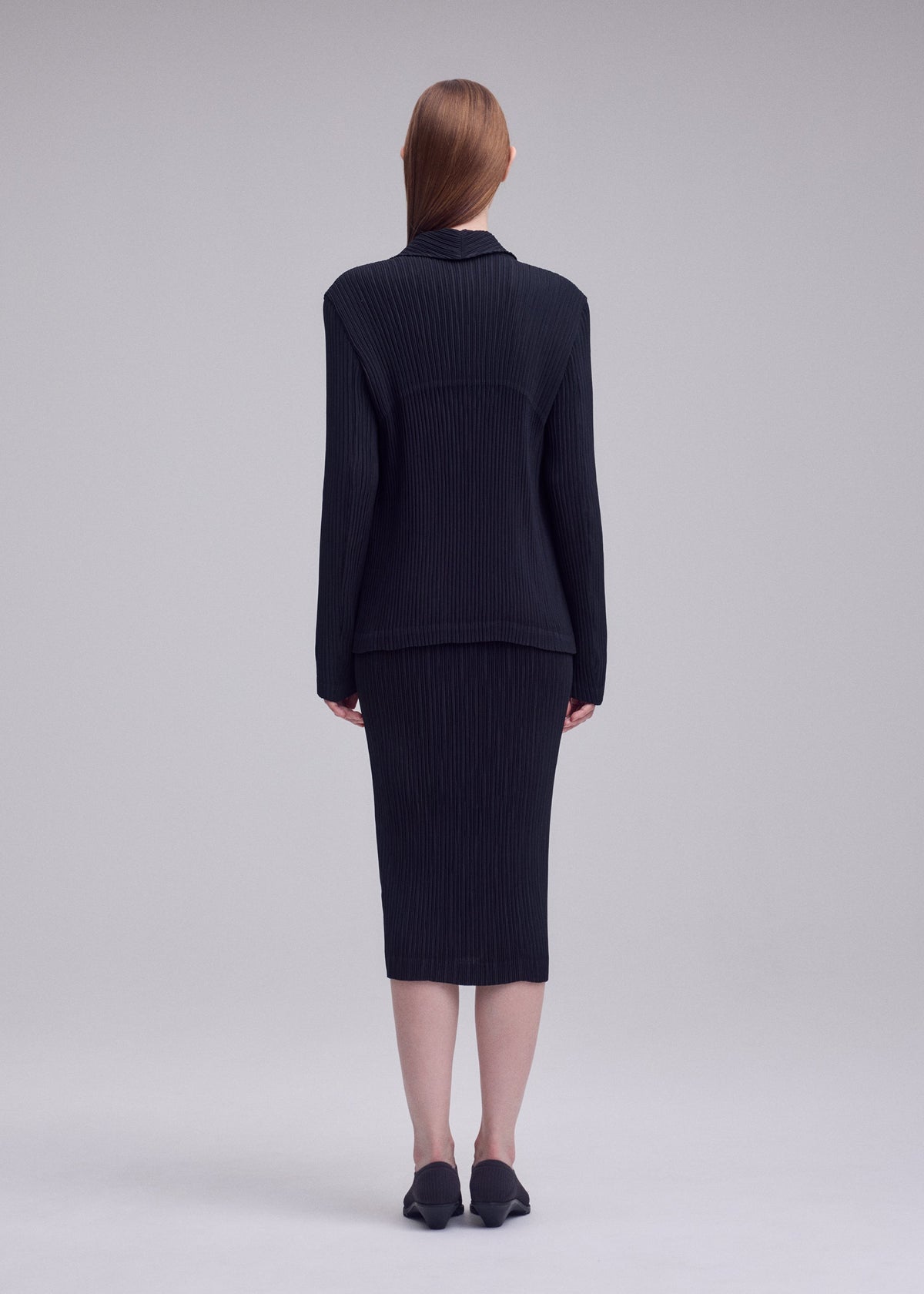 A person with straight, long hair stands facing away, wearing the HATCHING PLEATS set by ISSEY MIYAKE—black ribbed long-sleeve top and matching knee-length skirt—with black shoes against a light gray background. view 5 of 6