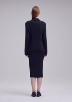 A person with straight, long hair stands facing away, wearing the HATCHING PLEATS set by ISSEY MIYAKE—black ribbed long-sleeve top and matching knee-length skirt—with black shoes against a light gray background.