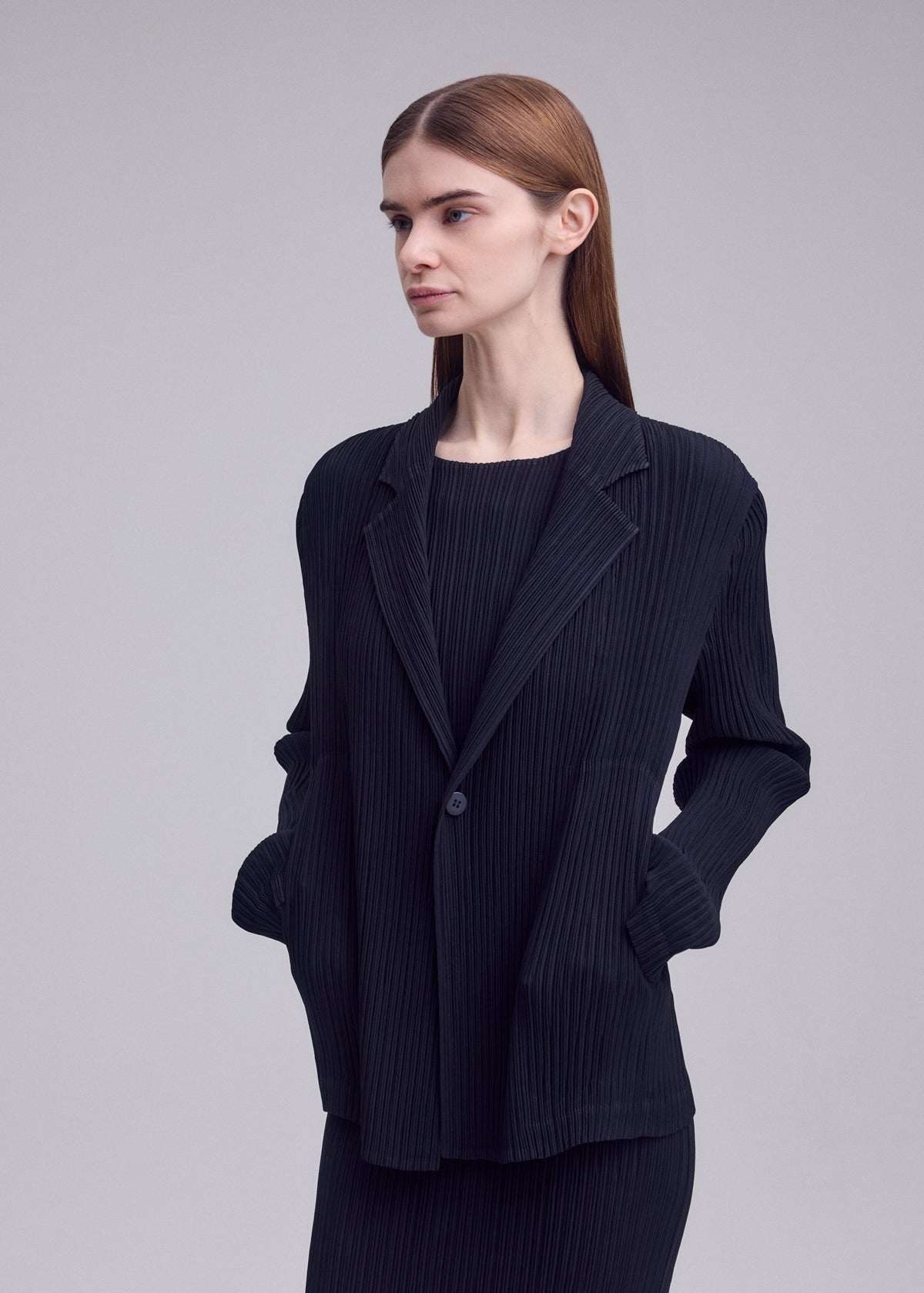 A person with straight brown hair wears the ISSEY MIYAKE HATCHING PLEATS black pinstripe tailored jacket, top, and skirt set, standing with hands in pockets against a plain light gray background. view 6 of 6