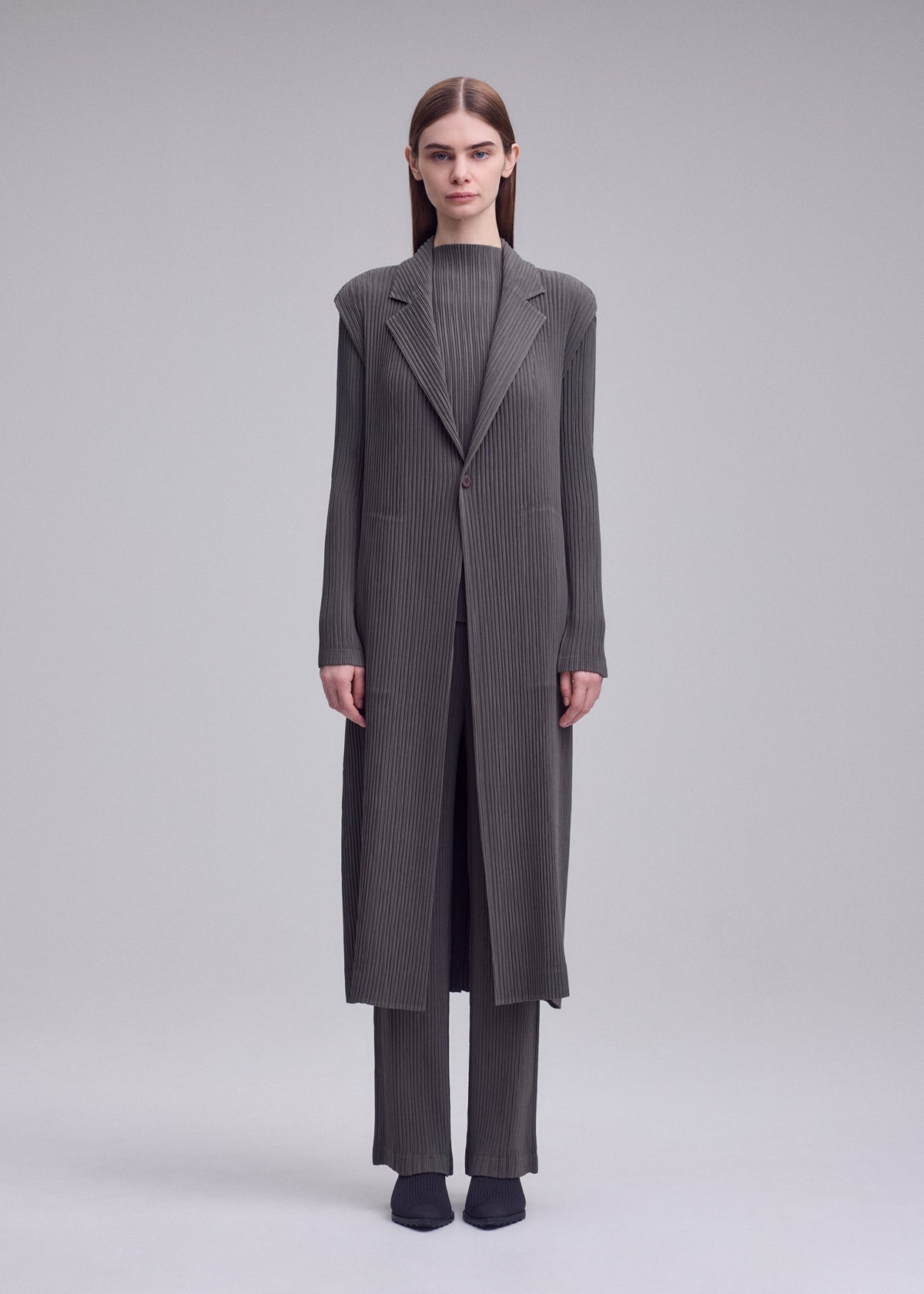 A woman faces forward in the ISSEY MIYAKE HATCHING PLEATS long gray pinstriped coat over matching pants and black shoes, set against a plain light gray background. view 4 of 6