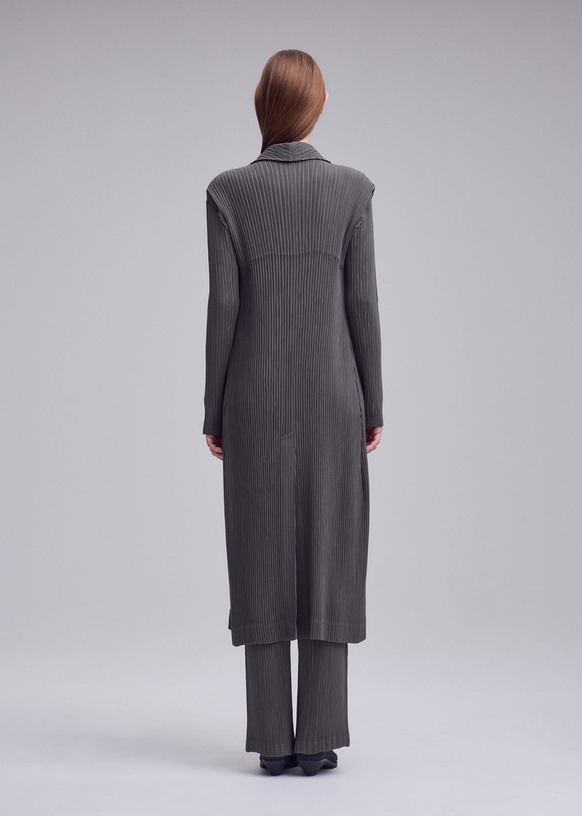 A person stands facing away, wearing ISSEY MIYAKE’s HATCHING PLEATS long vest and wide-leg pants in dark gray with a textured finish, paired with black shoes against a plain gray background. view 4 of 5