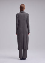 A person stands facing away, wearing ISSEY MIYAKE’s HATCHING PLEATS long vest and wide-leg pants in dark gray with a textured finish, paired with black shoes against a plain gray background.