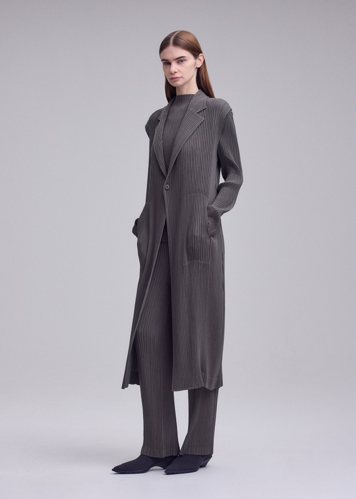 A woman poses against a plain background, wearing the ISSEY MIYAKE HATCHING PLEATS long gray pinstriped coat, layered over matching wide-leg pants and top. Her hands are in her pockets and her brown hair is tucked behind her ears. view 6 of 6