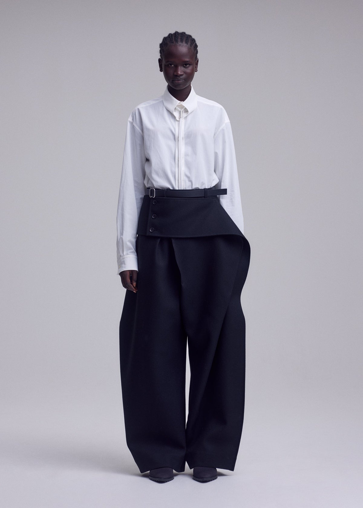 A person stands against a plain background, wearing a crisp white button-up shirt and high-waisted, wide-leg black CALLIGRAPH pants by ISSEY MIYAKE, featuring premium wool fabric and an asymmetrical belt detail. Their hair is short and styled, with a neutral expression. view 6 of 9