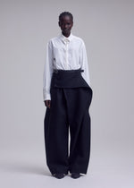 A person stands against a plain background, wearing a crisp white button-up shirt and high-waisted, wide-leg black CALLIGRAPH pants by ISSEY MIYAKE, featuring premium wool fabric and an asymmetrical belt detail. Their hair is short and styled, with a neutral expression.