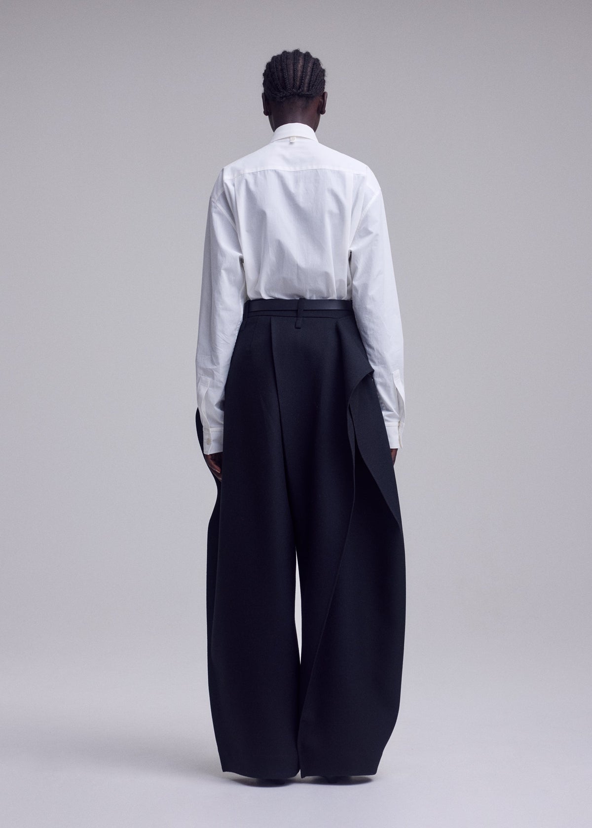 A person stands against a plain backdrop with their back to the camera, wearing a white button-up shirt paired with the ISSEY MIYAKE CALLIGRAPH high-waisted, wide-leg black trousers featuring draped side panels in premium woven nashiji wool, evoking the brand’s signature style. Their hair is styled in short braids. view 7 of 9