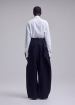 A person stands against a plain backdrop with their back to the camera, wearing a white button-up shirt paired with the ISSEY MIYAKE CALLIGRAPH high-waisted, wide-leg black trousers featuring draped side panels in premium woven nashiji wool, evoking the brand’s signature style. Their hair is styled in short braids.