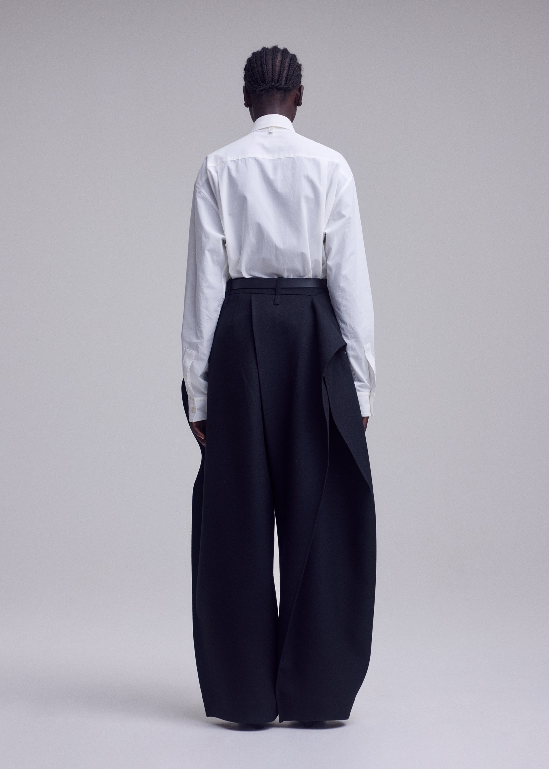Zoomed view A person stands against a plain backdrop with their back to the camera, wearing a white button-up shirt paired with the ISSEY MIYAKE CALLIGRAPH high-waisted, wide-leg black trousers featuring draped side panels in premium woven nashiji wool, evoking the brand’s signature style. Their hair is styled in short braids.
