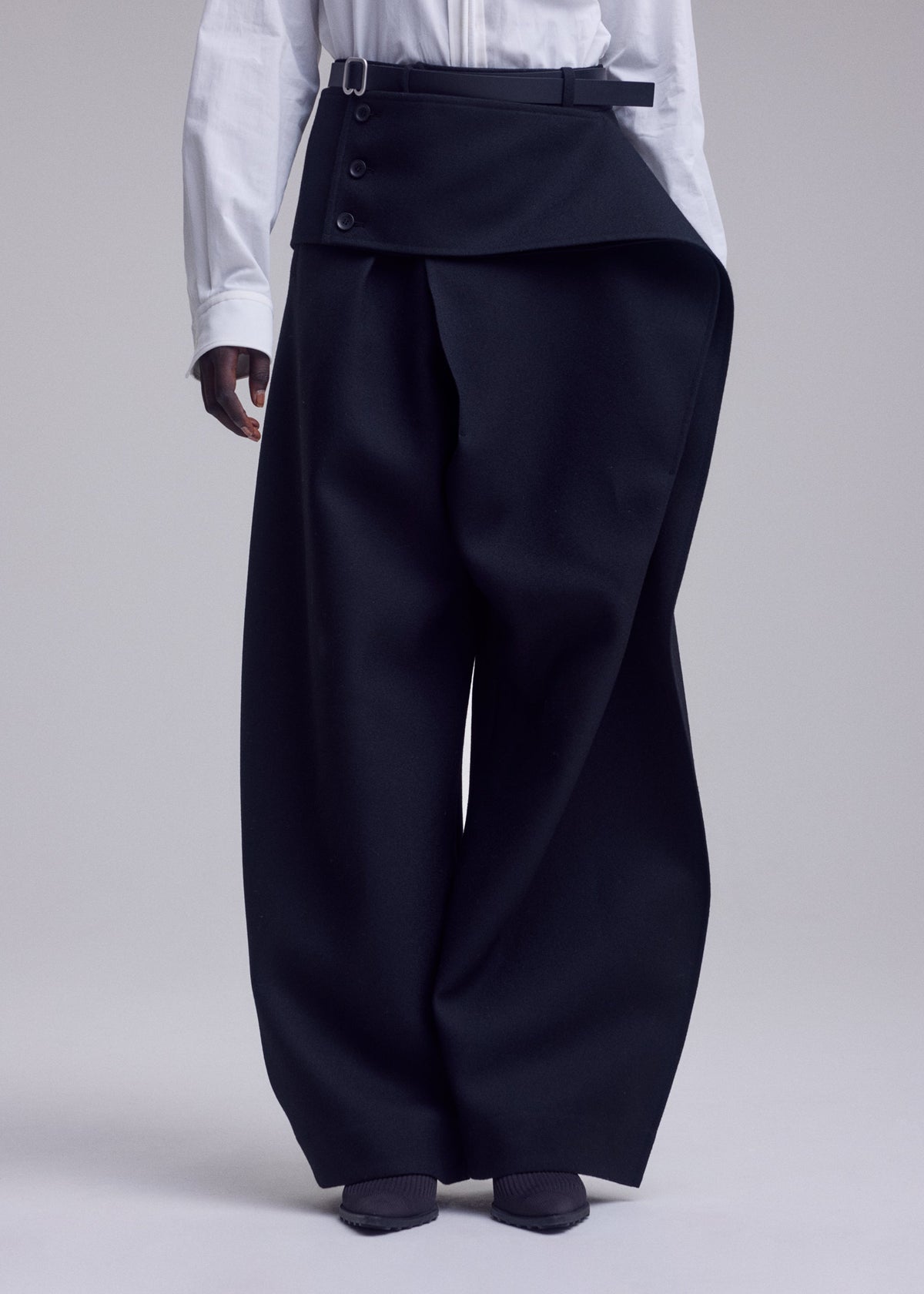 A person wearing a white shirt and ISSEY MIYAKE CALLIGRAPH wide-leg, high-waisted black premium wool pants featuring unique overlapping asymmetrical flap and button details, standing against a plain light background. Only the lower half of the body is visible. view 8 of 9
