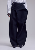A person wearing a white shirt and ISSEY MIYAKE CALLIGRAPH wide-leg, high-waisted black premium wool pants featuring unique overlapping asymmetrical flap and button details, standing against a plain light background. Only the lower half of the body is visible.