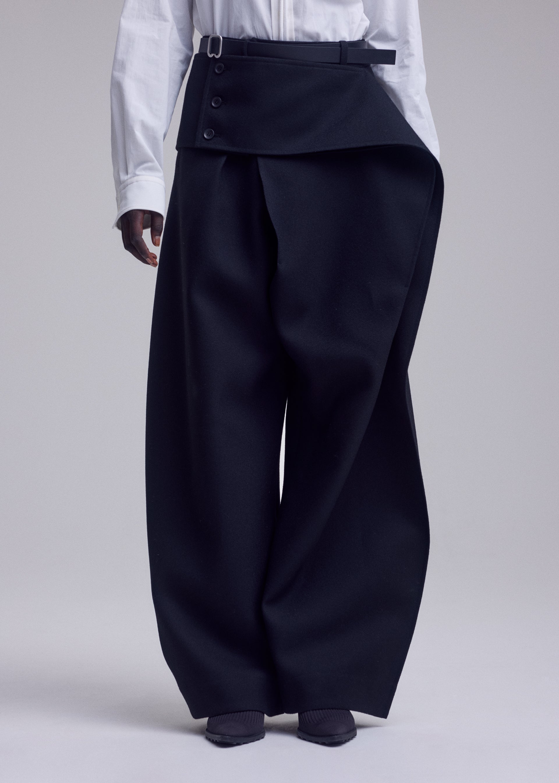 Zoomed view A person wearing a white shirt and ISSEY MIYAKE CALLIGRAPH wide-leg, high-waisted black premium wool pants featuring unique overlapping asymmetrical flap and button details, standing against a plain light background. Only the lower half of the body is visible.