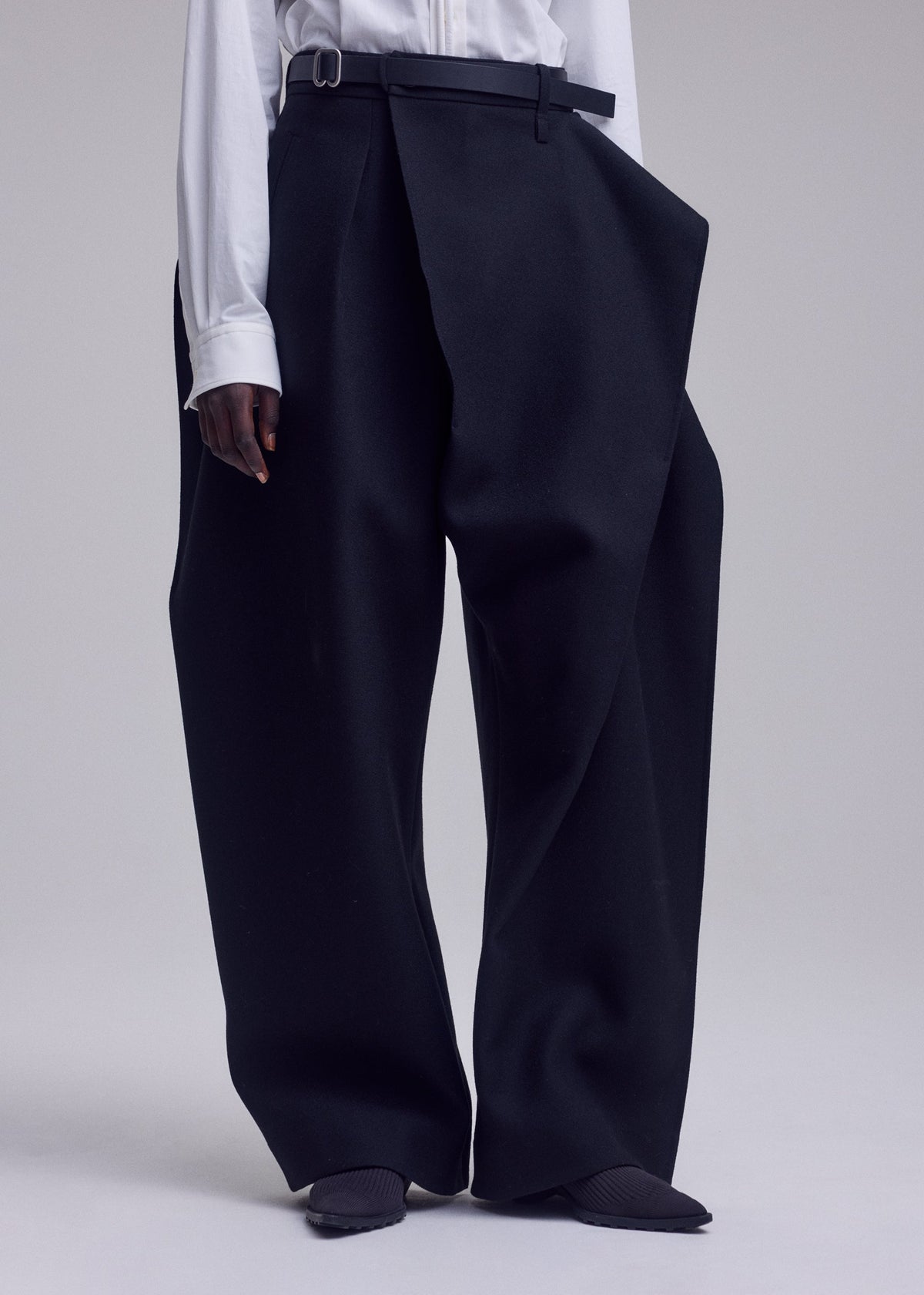 A person stands against a plain light background wearing the ISSEY MIYAKE CALLIGRAPH wide-leg black premium wool pants, which feature an asymmetrical wrap detail. The trousers, crafted from woven nashiji fabric for subtle texture and sophistication, are paired with a tucked-in white shirt and black pointed shoes. view 9 of 9