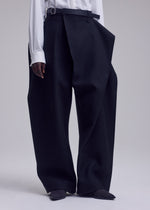 A person stands against a plain light background wearing the ISSEY MIYAKE CALLIGRAPH wide-leg black premium wool pants, which feature an asymmetrical wrap detail. The trousers, crafted from woven nashiji fabric for subtle texture and sophistication, are paired with a tucked-in white shirt and black pointed shoes.