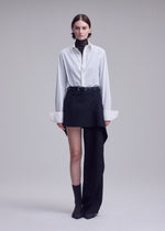 A person stands against a plain, light background in a look reminiscent of ISSEY MIYAKE aesthetics, featuring the CALLIGRAPH collection: a white button-up shirt layered over a dark turtleneck, paired with an asymmetrical black skirt uniquely combined with a single black premium wool pant leg—a striking display of asymmetric design—styled with gray socks and dark shoes.