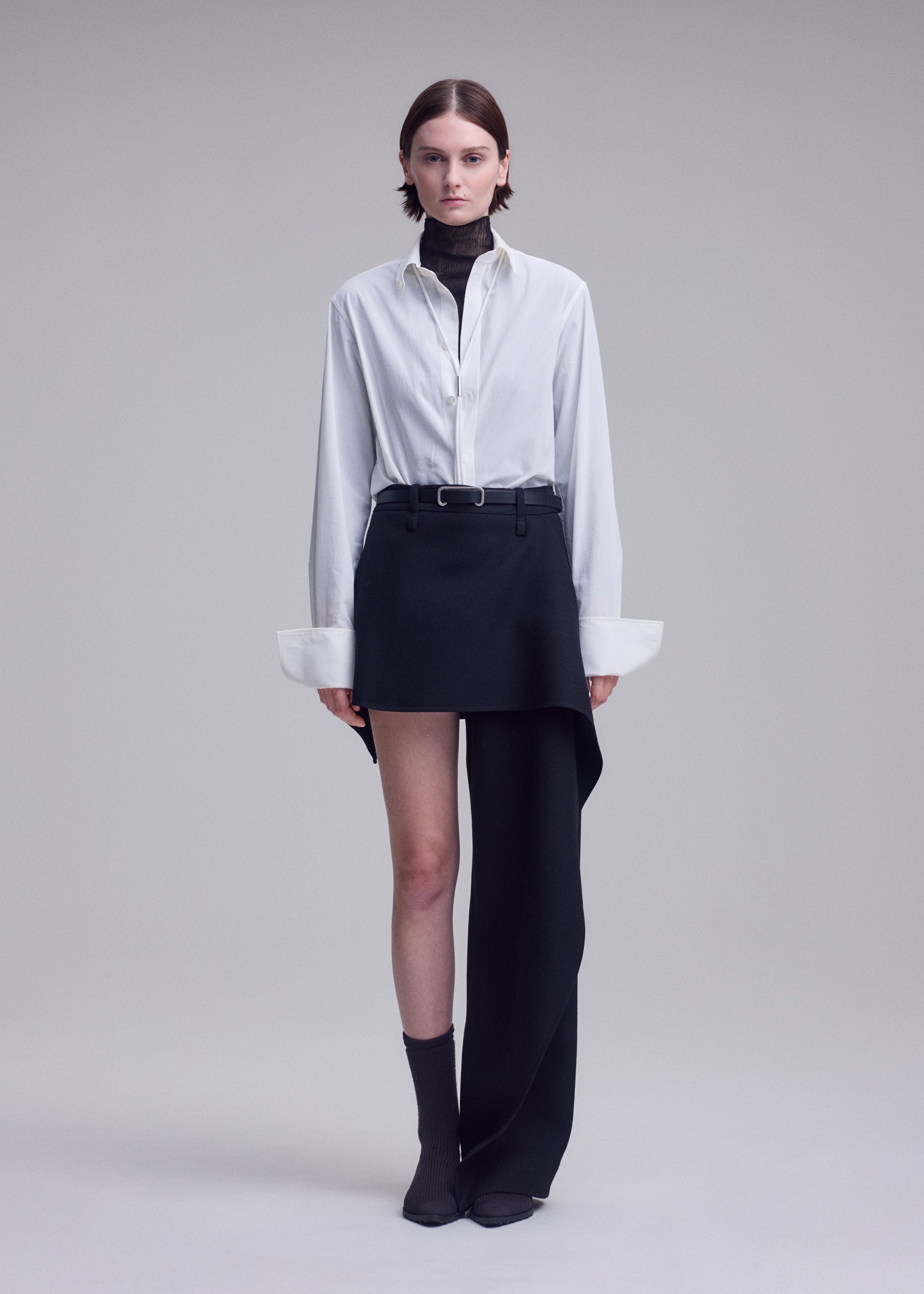 Zoomed view A person stands against a plain, light background in a look reminiscent of ISSEY MIYAKE aesthetics, featuring the CALLIGRAPH collection: a white button-up shirt layered over a dark turtleneck, paired with an asymmetrical black skirt uniquely combined with a single black premium wool pant leg—a striking display of asymmetric design—styled with gray socks and dark shoes.
