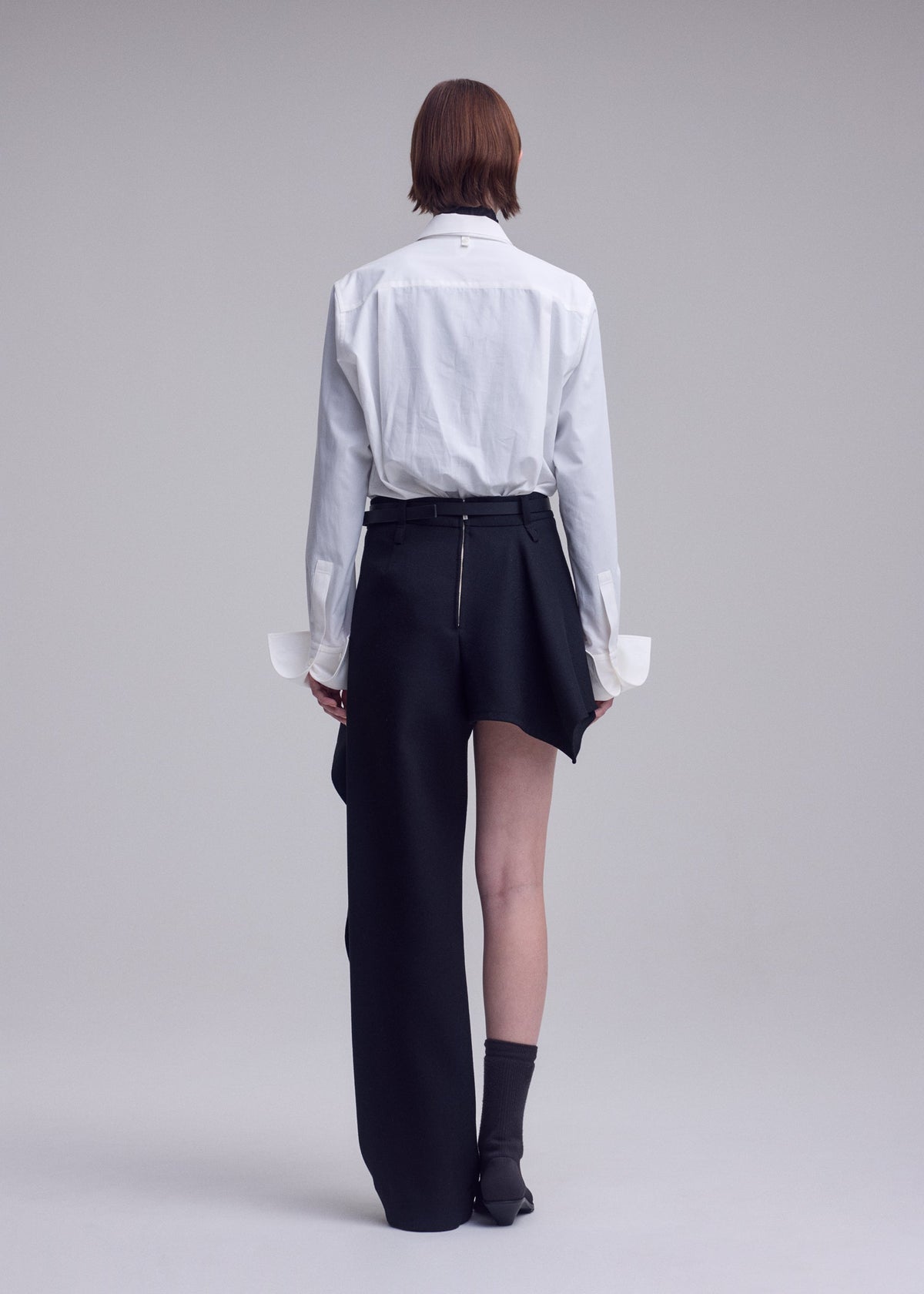 Standing against a plain background, the individual wears a white button-up shirt and ISSEY MIYAKE’s CALLIGRAPH skirt-pants, which feature an asymmetric design with one leg covered while the other remains bare, paired with black socks and shoes; their back is turned to the camera. view 7 of 8