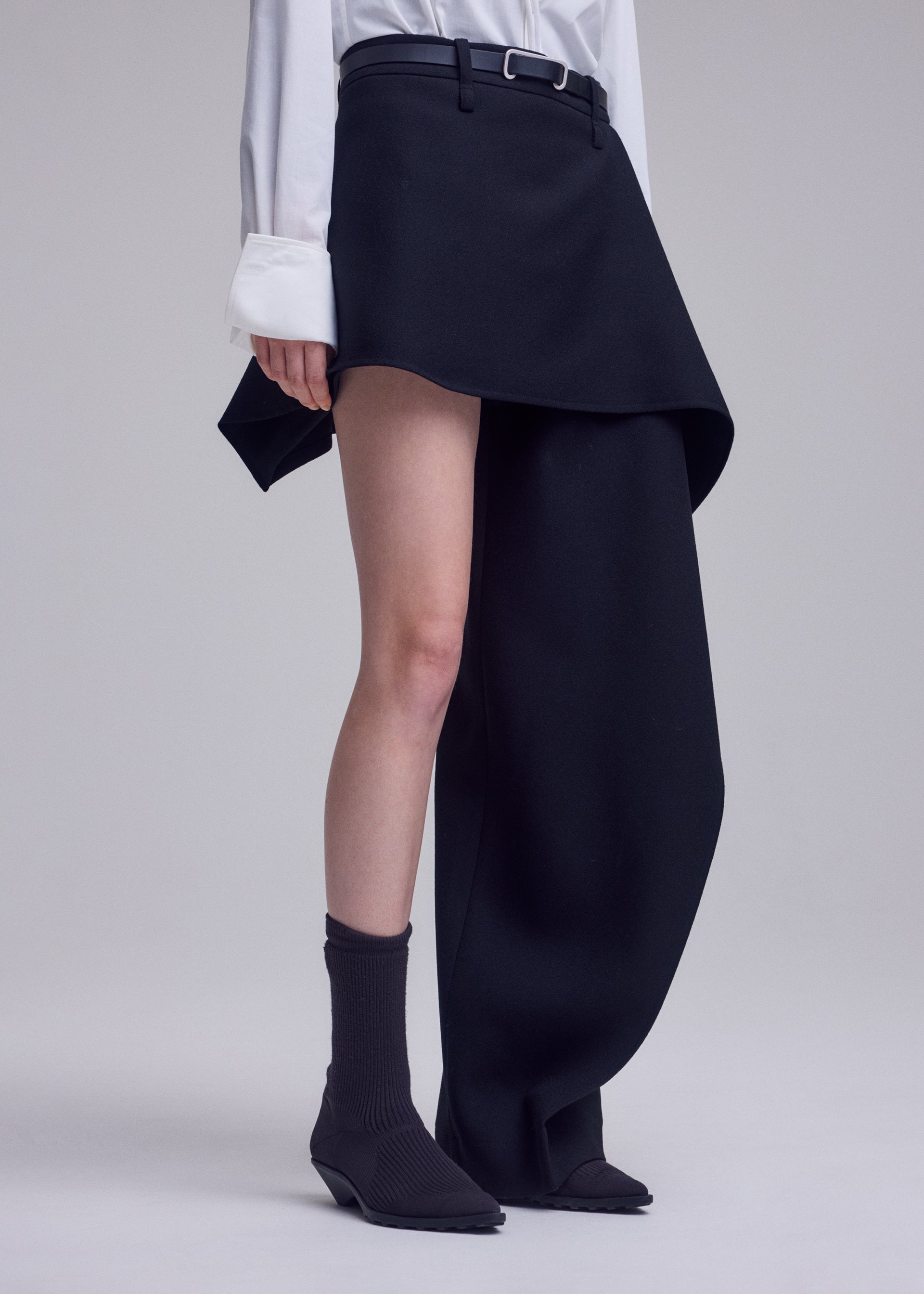 Zoomed view A person wearing a white shirt, the ISSEY MIYAKE CALLIGRAPH asymmetrical black skirt, black ankle socks, and black shoes stands against a plain background.