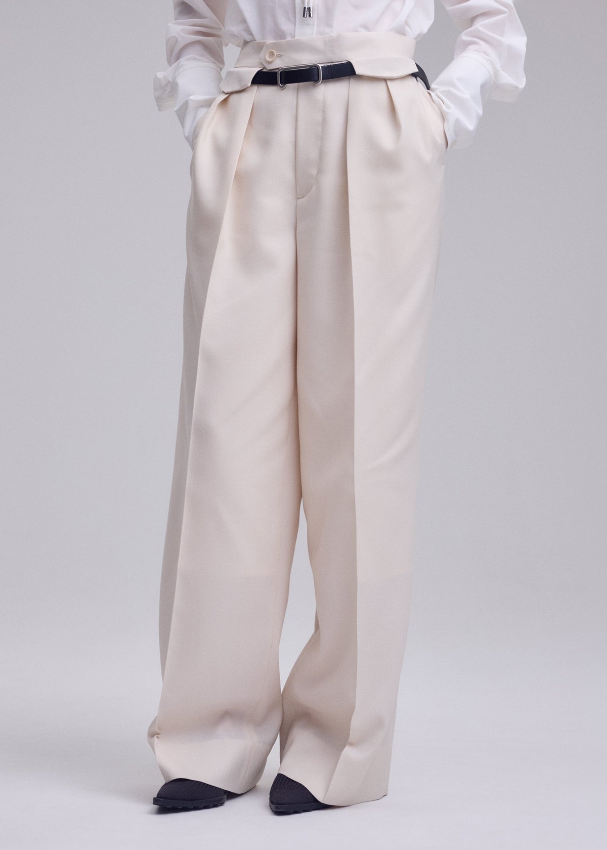 Zoomed view Person wearing UNTITLED wool wide pants by ISSEY MIYAKE in cream, styled with a black belt and tucked-in white shirt. Hands in pockets; only the lower body is shown against a plain background.