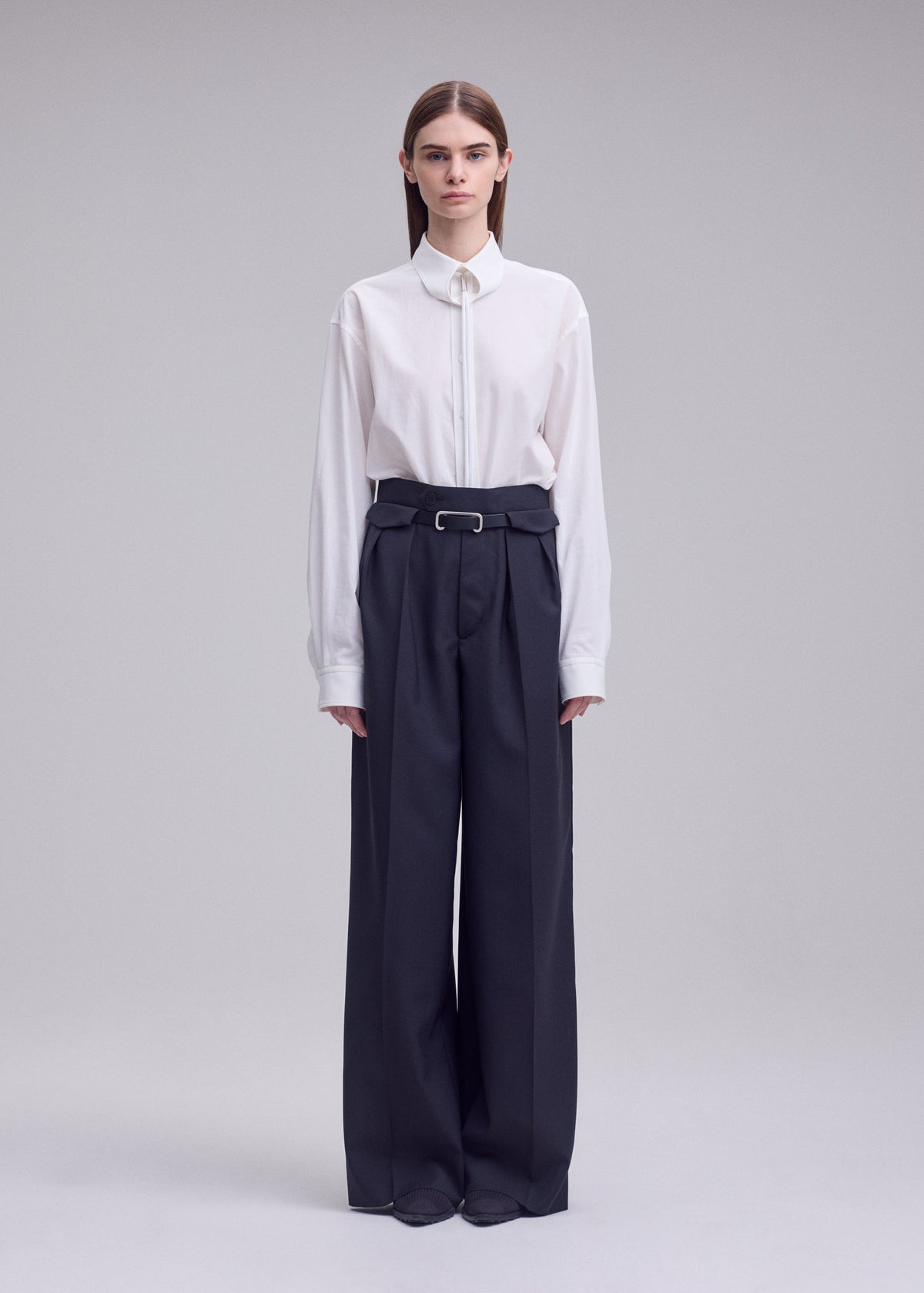 A person stands against a plain background, wearing the HONEST ENSEMBLE by ISSEY MIYAKE: a white collared shirt tucked into high-waisted, wide yarn-dyed wool pants with a belt, and has straight hair parted down the middle. view 5 of 7