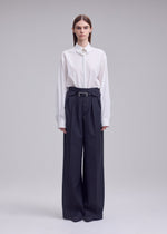 A person stands against a plain background, wearing the HONEST ENSEMBLE by ISSEY MIYAKE: a white collared shirt tucked into high-waisted, wide yarn-dyed wool pants with a belt, and has straight hair parted down the middle.