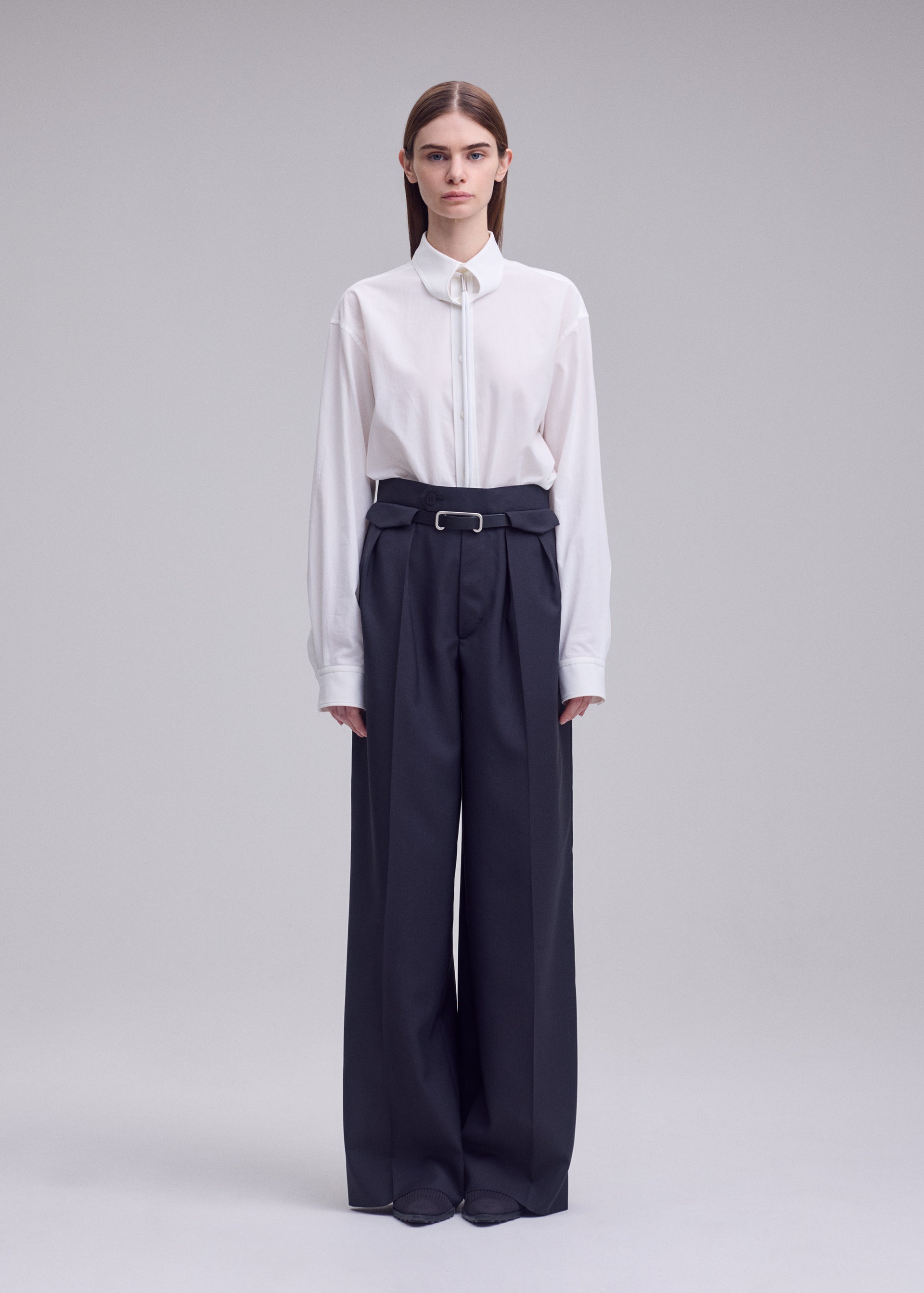 Zoomed view A person stands against a plain background, wearing the HONEST ENSEMBLE by ISSEY MIYAKE: a white collared shirt tucked into high-waisted, wide yarn-dyed wool pants with a belt, and has straight hair parted down the middle.