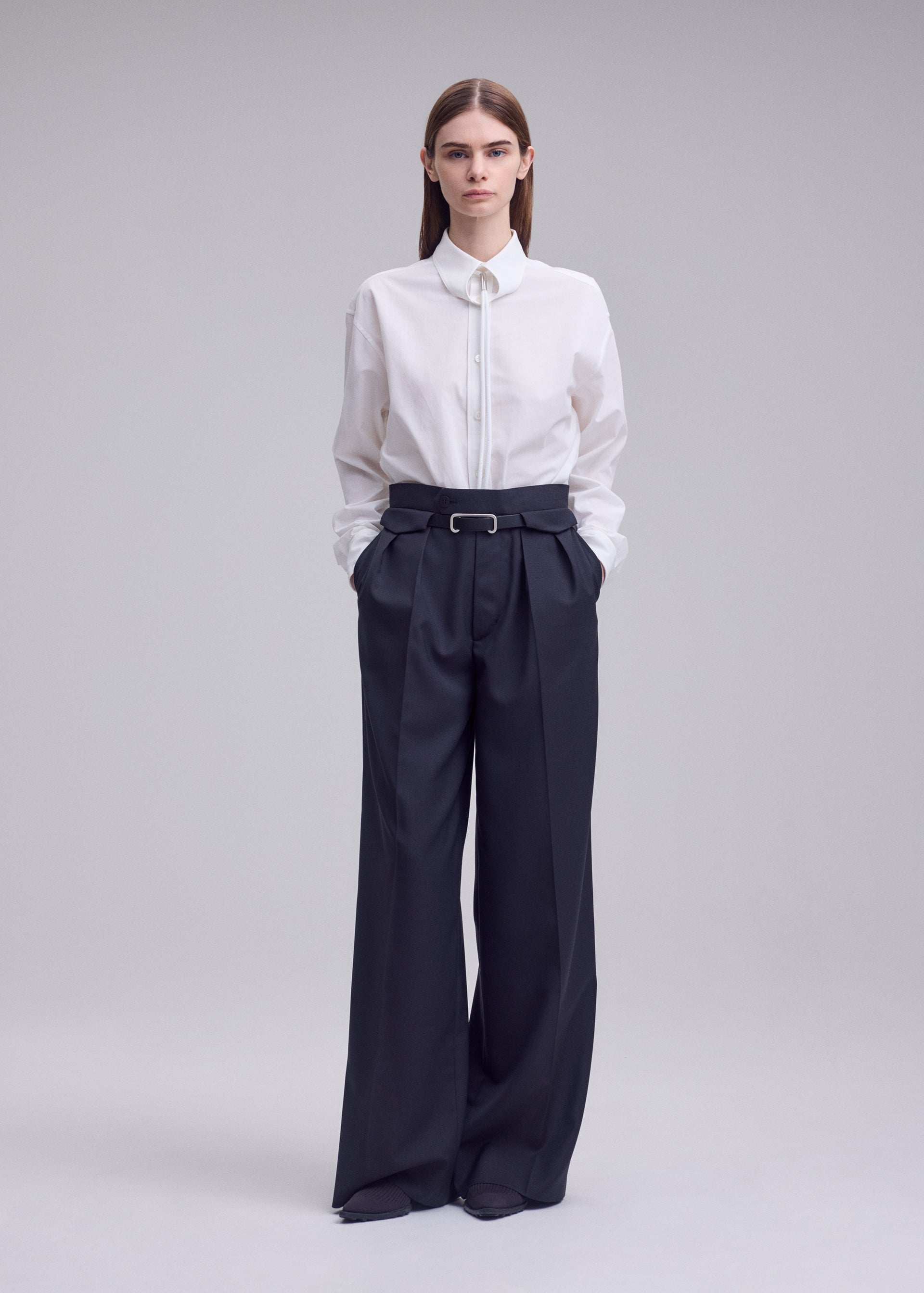 Zoomed view A person stands facing forward against a plain light background, wearing the HONEST ENSEMBLE by ISSEY MIYAKE—a white button-up shirt tucked into high-waisted, wide-leg pants crafted from yarn-dyed wool with a belt—hands in pockets.