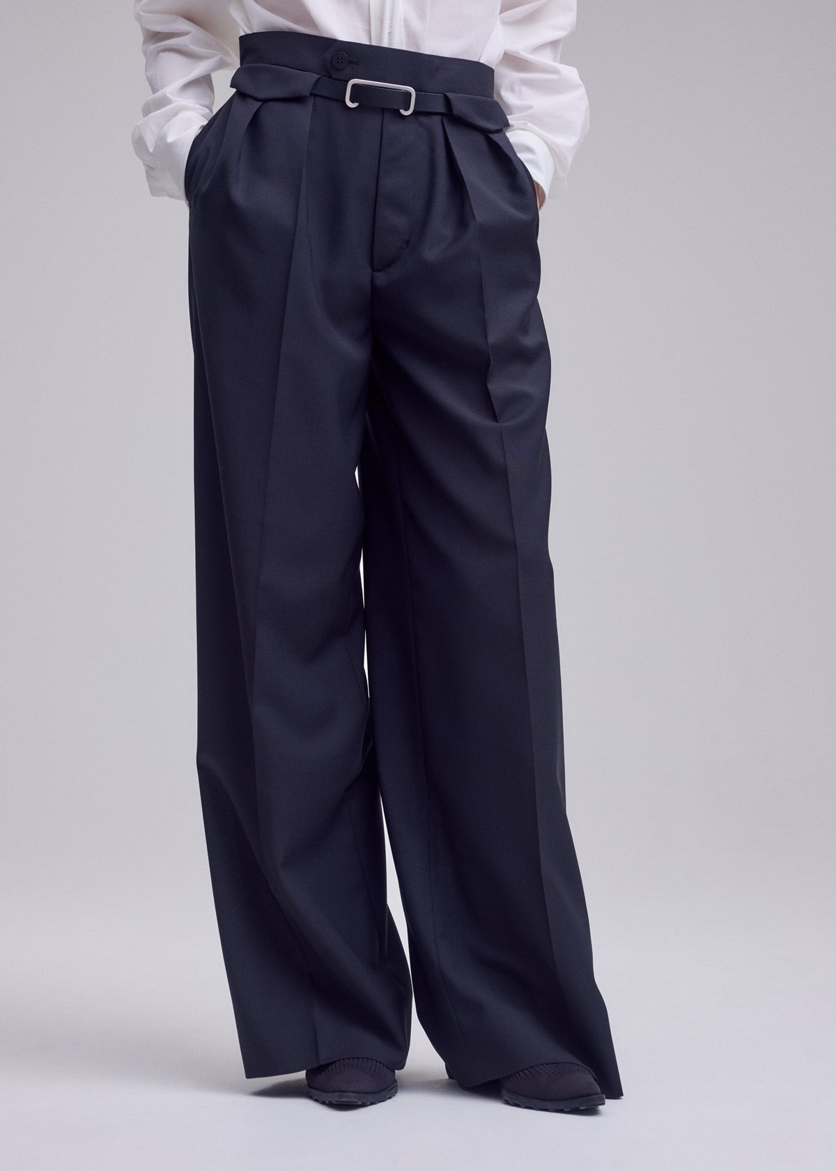 A person is shown from the waist down, dressed in the HONEST ENSEMBLE wide-leg, high-waisted black trousers by ISSEY MIYAKE, expertly tailored from premium yarn-dyed wool and distinguished by elegant pleats, belt loops, and a silver bar detail at the waist. The look is completed with a crisp white shirt. view 7 of 7