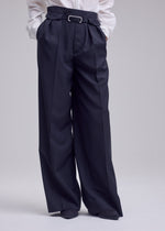 A person is shown from the waist down, dressed in the HONEST ENSEMBLE wide-leg, high-waisted black trousers by ISSEY MIYAKE, expertly tailored from premium yarn-dyed wool and distinguished by elegant pleats, belt loops, and a silver bar detail at the waist. The look is completed with a crisp white shirt.