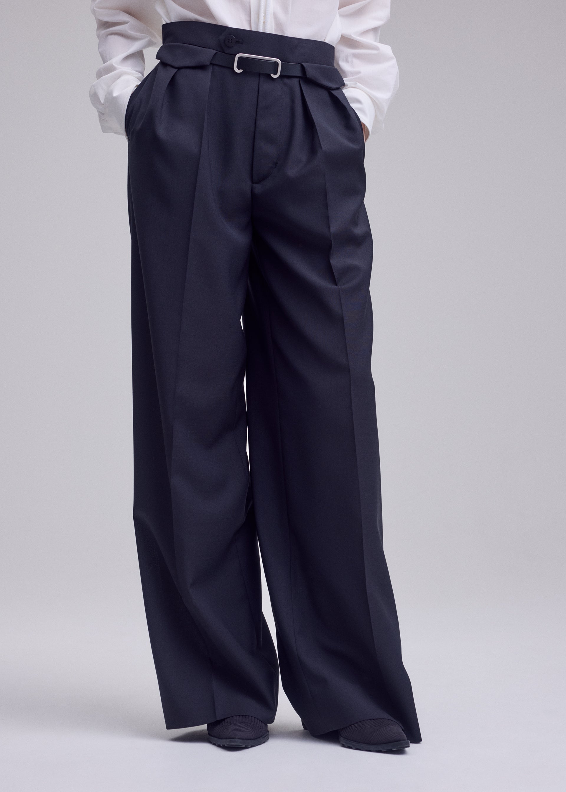 Zoomed view A person is shown from the waist down, dressed in the HONEST ENSEMBLE wide-leg, high-waisted black trousers by ISSEY MIYAKE, expertly tailored from premium yarn-dyed wool and distinguished by elegant pleats, belt loops, and a silver bar detail at the waist. The look is completed with a crisp white shirt.