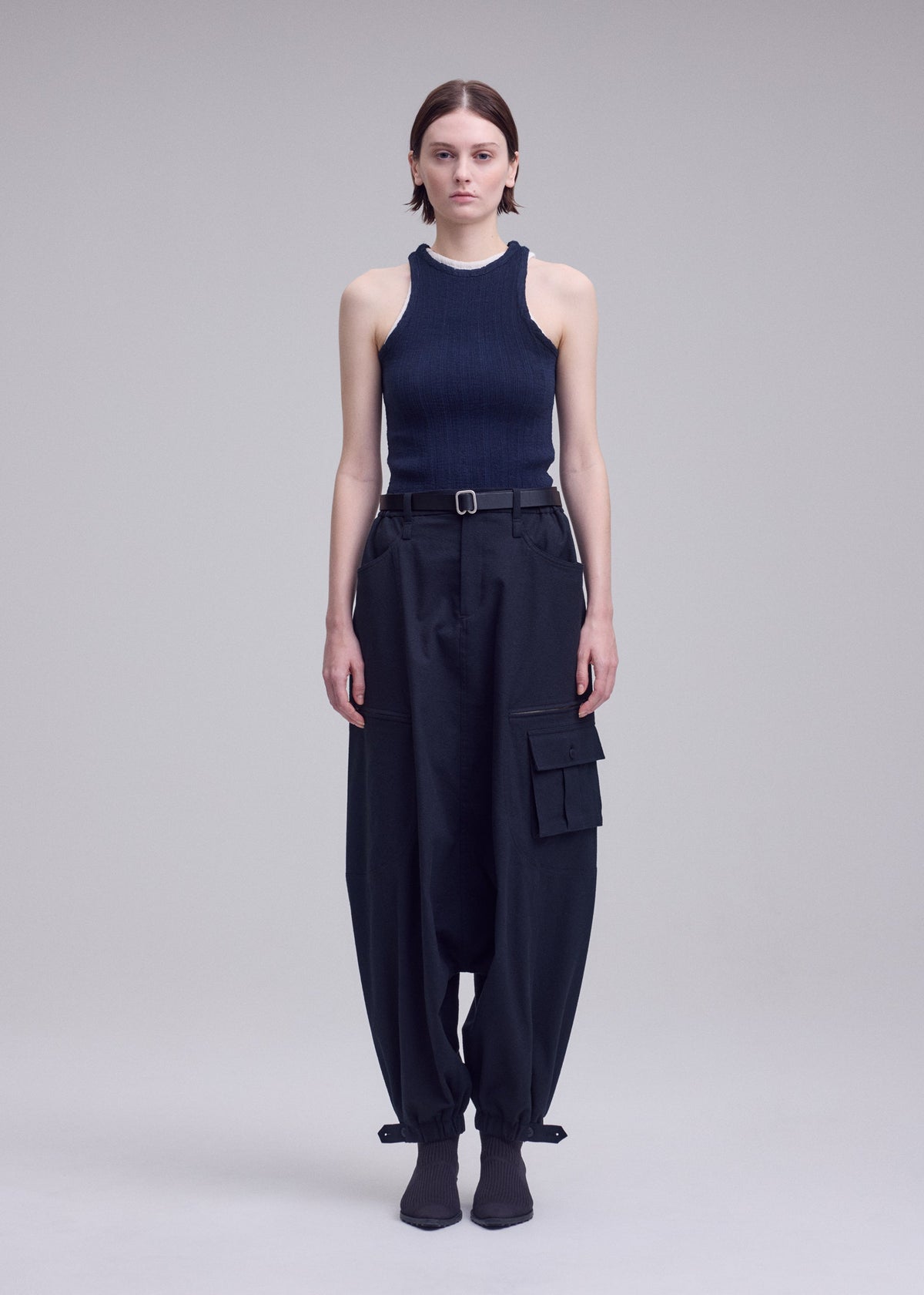 A person with straight brown hair stands against a plain background, wearing a sleeveless black ribbed top, ISSEY MIYAKE GRAINY PANTS featuring jacquard weaving and large pockets, styled with a belt and heeled boots. view 6 of 8