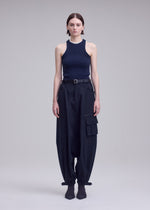 A person with straight brown hair stands against a plain background, wearing a sleeveless black ribbed top, ISSEY MIYAKE GRAINY PANTS featuring jacquard weaving and large pockets, styled with a belt and heeled boots.